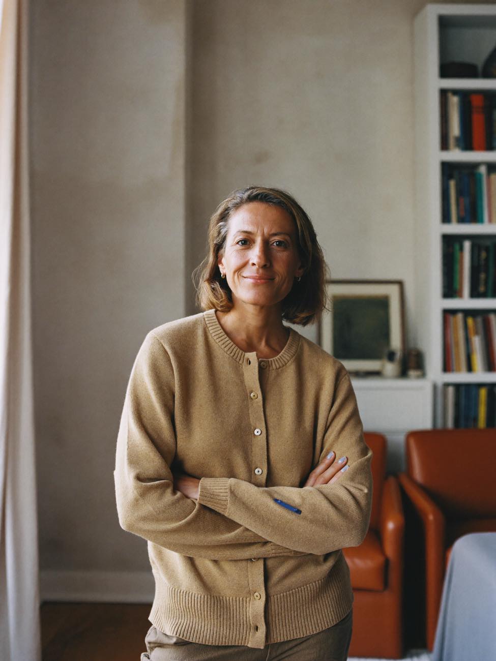 A woman in a camel cardigan stands with her arms folded in a bright living room lined with bookshelves, smiling at the camera.